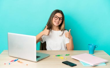 Masada dizüstü bilgisayarı olan küçük bir kız izole edilmiş mavi arka planda baş parmak işareti yapıyor.