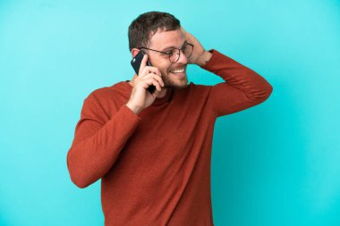 Mavi arka planda izole edilmiş cep telefonu kullanan genç Brezilyalı bir adam elini kulağına koyarak bir şeyler dinliyor.