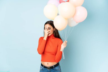 Mavi arka planda izole edilmiş balonları yakalayan genç bir kadın gözleri kapatıyor ve parmaklarından bakıyor.