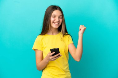 Küçük kız izole edilmiş mavi arka planda, telefon zafer pozisyonunda.