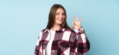 Genç beyaz kız mavi arka planda izole edilmiş. Parmaklarıyla tamam işareti gösteriyor.