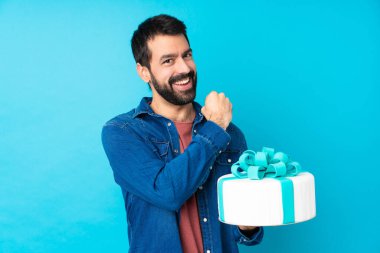 Büyük pastası olan genç yakışıklı adam izole edilmiş mavi arka planda zaferi kutluyor.