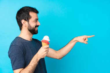 Kornet dondurması olan genç bir adam, izole edilmiş mavi arka planda parmak göstererek bir ürün sunuyor.