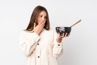 Beyaz arka planda, elinde yemek çubuklarıyla elinde bir kase şehriye tutarken, şaşırmış ve şok olmuş bir yüz ifadesi olan beyaz bir kız.