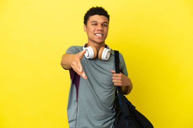 Sarı arka planda izole edilmiş spor çantalı genç Afrikalı Amerikalı adam iyi bir anlaşma için el sıkışıyor.