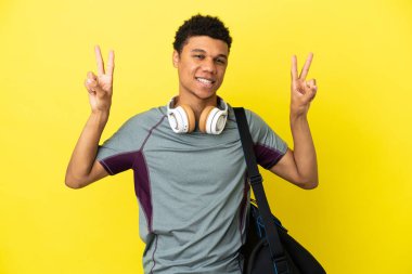 Sarı arka planda izole edilmiş spor çantalı genç Afrikalı Amerikalı iki eliyle zafer işareti gösteriyor.