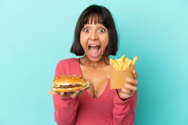Genç esmer kadın, elinde hamburger ve kızarmış cipslerle izole edilmiş mavi arka planda.