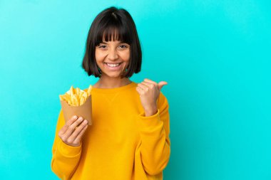 İzole edilmiş mavi arka planda patates kızartması tutan genç esmer kadın bir ürünü sunmak için kenarı işaret ediyor.