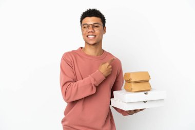 Elinde hamburgerle beyaz arka planda izole edilmiş pizzalar tutan genç Afrikalı Amerikalı bir adam bir ürün sunmak için kenarı işaret ediyor.