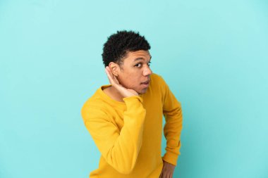 Genç Afrikalı Amerikalı bir adam mavi arka planda izole edilmiş. Kulağına elini sürerek bir şeyler dinliyor.