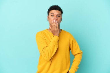 Genç Afrikalı Amerikalı bir adam mavi arka planda izole edilmiş, ağzı elle kapatılmış.