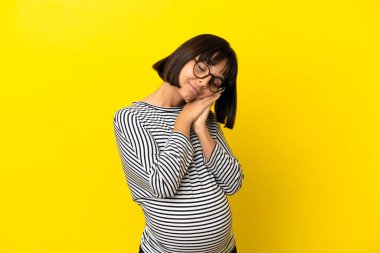 İzole edilmiş sarı arka planda genç hamile bir kadın uyku jesti yapıyor.