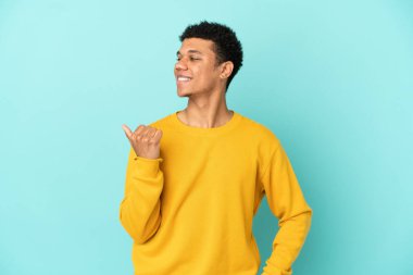 Mavi arka planda izole edilmiş genç Afrikalı Amerikalı bir ürün sunmak için kenarı işaret ediyor.