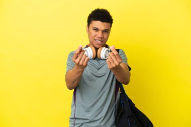 Sarı arka planda izole edilmiş spor çantalı genç Afro-Amerikalı adam para hareketi yapıyor.