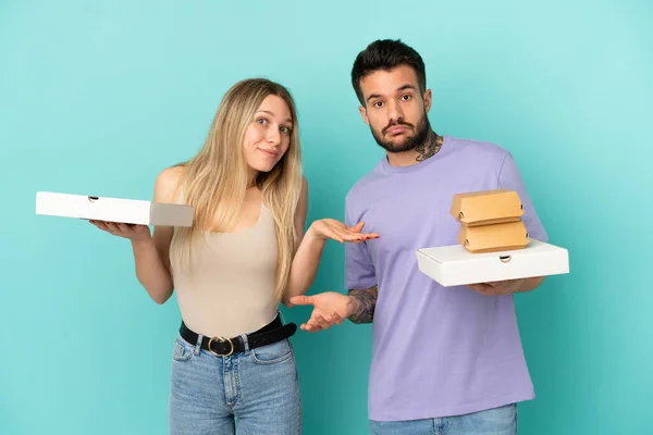 Casal Segurando Pizzas Hambúrgueres Sobre Fundo Azul Isolado Ter Dúvidas — Fotografia de Stock