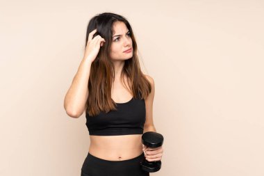 Genç bayan halterci bej arka planda izole haldeyken kuşkuları ve kafa karıştırıcı yüz ifadesi ile halter kaldırıyor