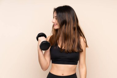 Genç bayan halter yapıyor. Bej arka planda izole edilmiş. Yan tarafa bakıyor.