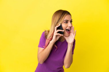 İzole edilmiş sarı arka planda cep telefonu kullanan genç bir kadın ağzı sonuna kadar açık bağırıyor.