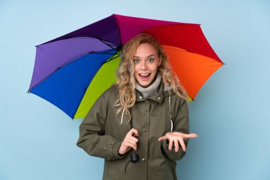 Mavi arka planda şemsiyesi olan genç sarışın kadın şok olmuş yüz ifadesi ile izole edilmiş.