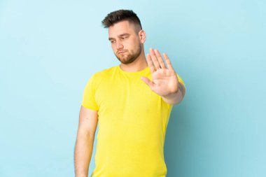 Rus yakışıklı adam mavi arka planda izole edilmiş. Durma hareketi yapıyor ve hayal kırıklığına uğruyor.