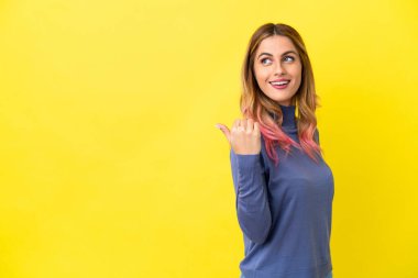 İzole edilmiş sarı arka planda genç bir kadın bir ürün sunmak için kenarı işaret ediyor.