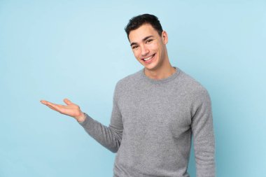 Beyaz tenli yakışıklı adam mavi arka planda izole edilmiş. Davet etmek için ellerini yana uzatıyor.