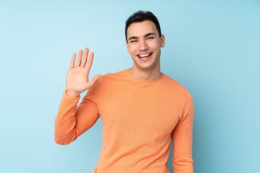 Beyaz tenli yakışıklı adam mavi arka planda izole edilmiş mutlu bir ifadeyle selamlıyor.