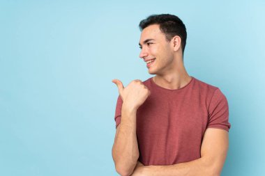 Mavi arka planda izole edilmiş beyaz, yakışıklı genç bir adam bir ürün sunmak için kenarı işaret ediyor.