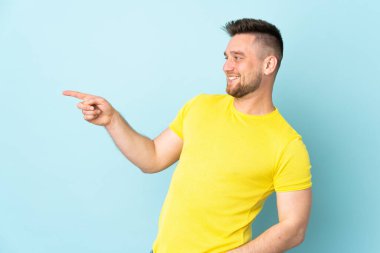 Rus yakışıklı bir adam mavi arka planda izole edilmiş, parmağını yana doğru uzatıyor ve bir ürün sunuyor.