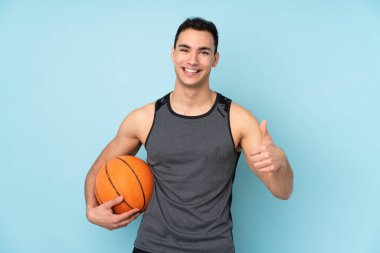 İzole edilmiş mavi arka planda basketbol oynayan ve başparmağı havada bir adam.