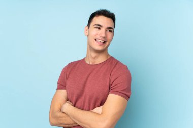 Mavi arka planda izole edilmiş, kolları bağlı ve mutlu, beyaz, yakışıklı genç bir adam.