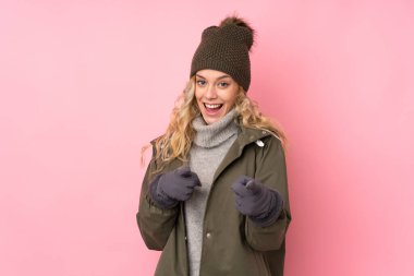 Kış şapkalı genç kız pembe arka planda izole edilmiş sana parmak atıyor.