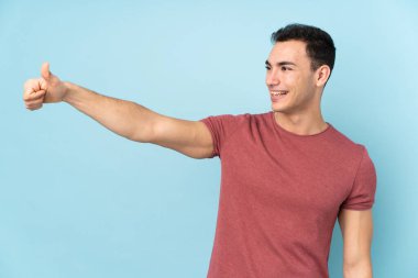 Beyaz tenli yakışıklı adam mavi arka planda izole edilmiş bir şekilde baş parmak işareti yapıyor.