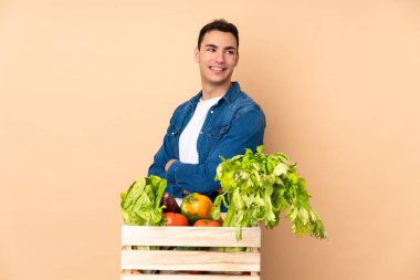 Bej arka planda izole edilmiş, kolları çapraz ve mutlu bir kutuda taze sebze toplayan çiftçi.