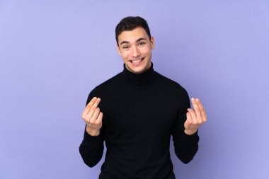 Balıkçı yaka kazaklı, mor arka planda izole edilmiş yakışıklı genç adam para hareketi yapıyor.