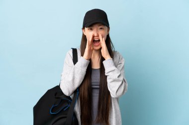 Spor çantalı genç bir Çinli kadın izole edilmiş mavi arka planda bağırıyor ve bir şeyler söylüyor.