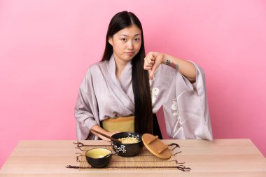 Kimono giyen ve erişte yiyen genç Çinli kız olumsuz bir ifadeyle baş parmağını aşağı gösteriyor.