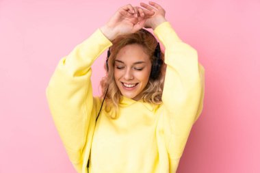 Pembe arka planda izole edilmiş sweatshirt giyen genç sarışın kadın müzik dinliyor ve dans ediyor.