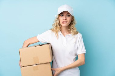 Mavi arka planda izole edilmiş genç teslimatçı kadının şüpheleri ve kafa karıştırıcı bir yüz ifadesi var.