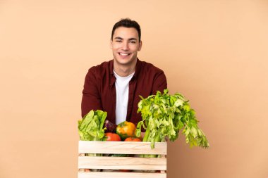 Bej arka planda izole bir kutuda taze toplanmış sebzelerle çiftçi kollarını ön konumda çapraz tutuyor.