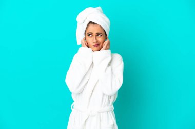 İzole edilmiş mavi arka planda bornozlu genç bir kadın hayal kırıklığına uğramış ve kulaklarını tıkamış.