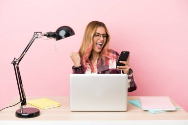 İş yerinde pembe arka planda dizüstü bilgisayarı olan ve telefonu zafer pozisyonunda olan genç bir bayan öğrenci.