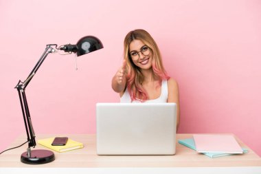 İş yerindeki pembe arka planda dizüstü bilgisayarı olan genç bir bayan iyi bir anlaşma yaptığı için el sıkışıyor.