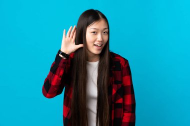 Genç Çinli kız mavi arka planda bir şeyler dinlerken elini kulağına koydu.