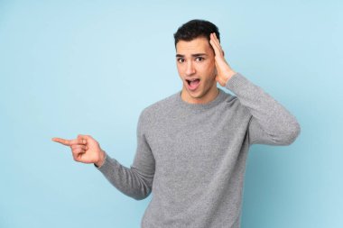 Mavi arka planda izole edilmiş, genç, beyaz, yakışıklı bir adam şaşırmış ve parmağıyla kenarı işaret ediyor.
