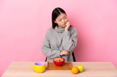 Genç Çinli kız yorgun ve sıkılmış bir ifadeyle bir masada kahvaltı yapıyor.