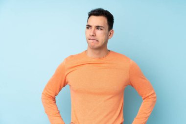 Kafkasyalı yakışıklı genç adam mavi arka planda izole edilmiş ve kafası karışmış bir yüz ifadesi var.