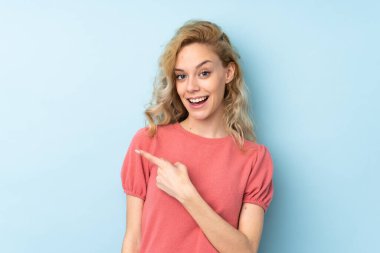 Mavi arka planda izole edilmiş genç sarışın kadın parmağını yana doğru gösteriyor.