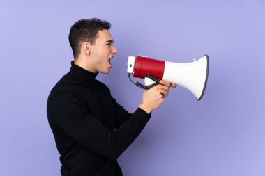 Balıkçı yaka kazaklı, genç beyaz bir adam mor arka planda izole edilmiş megafondan bağırıyor.