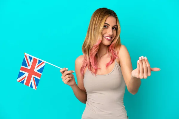 Young woman holding an United Kingdom flag over isolated blue background inviting to come with hand. Happy that you came